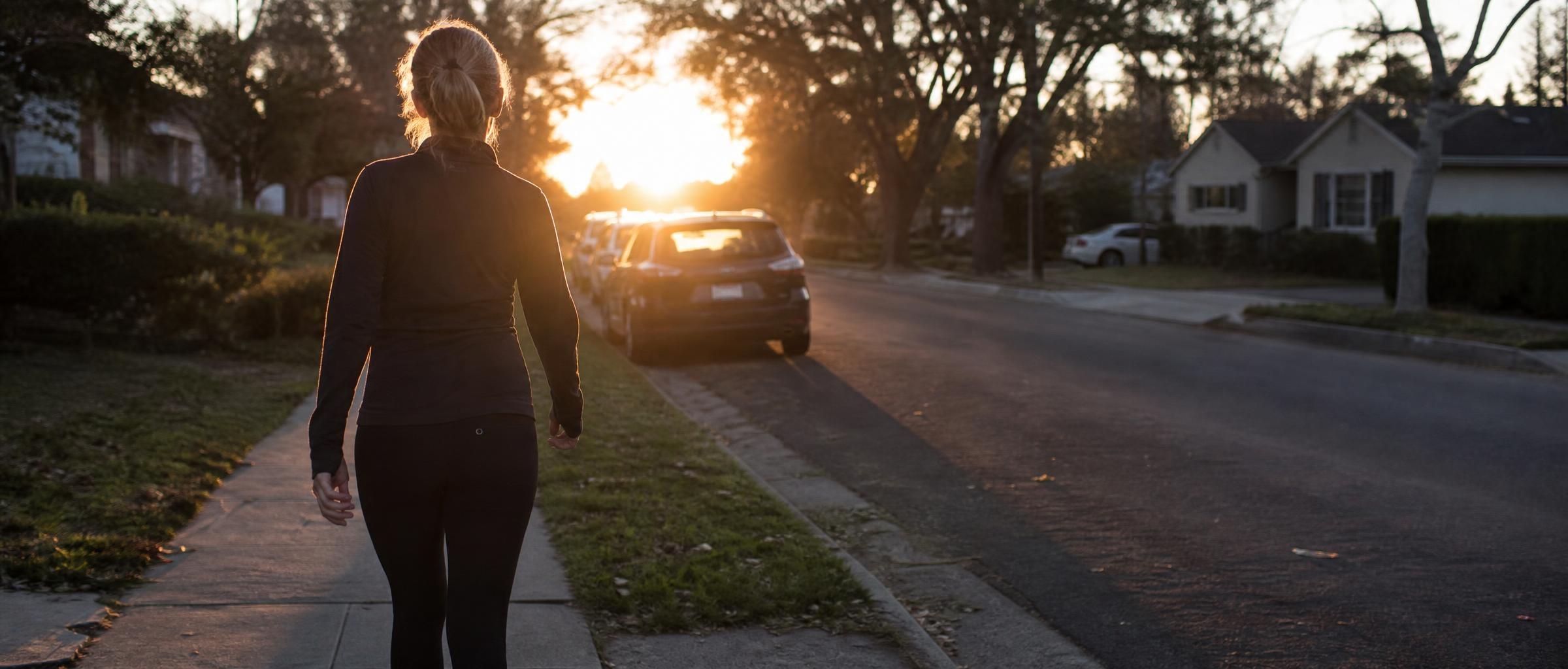 A woman in her mid-forties takes her first steps along a sidewalk at dawn, backlit by a low sun, her shadow stretching behind her.