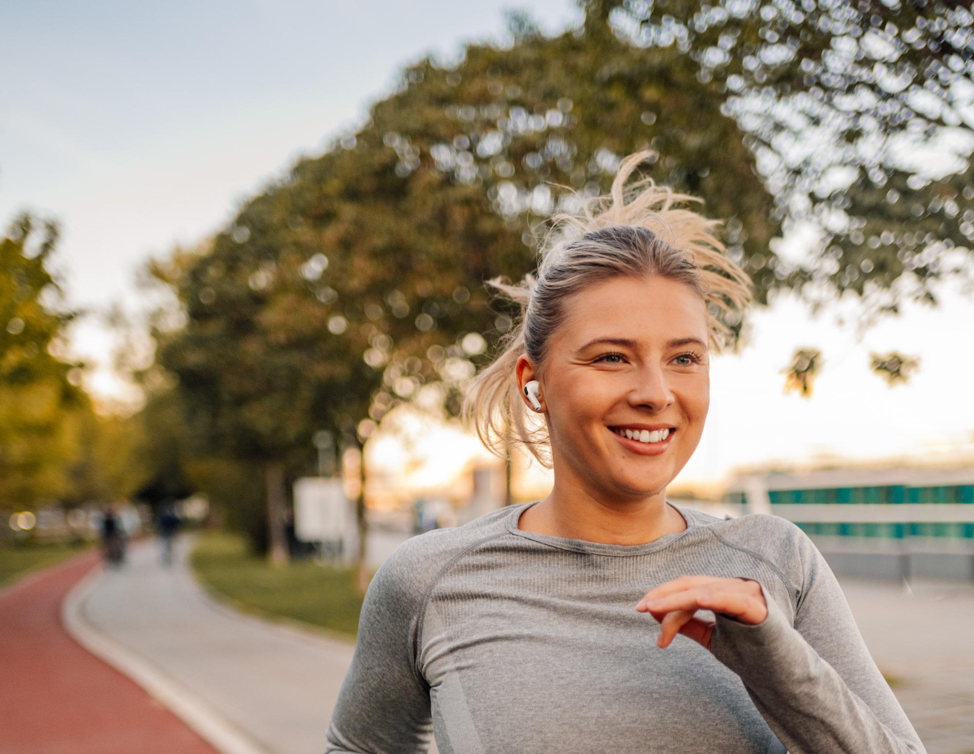 A smiling woman with a white earbud running toward the camera on a tree-lined park path in warm evening light.